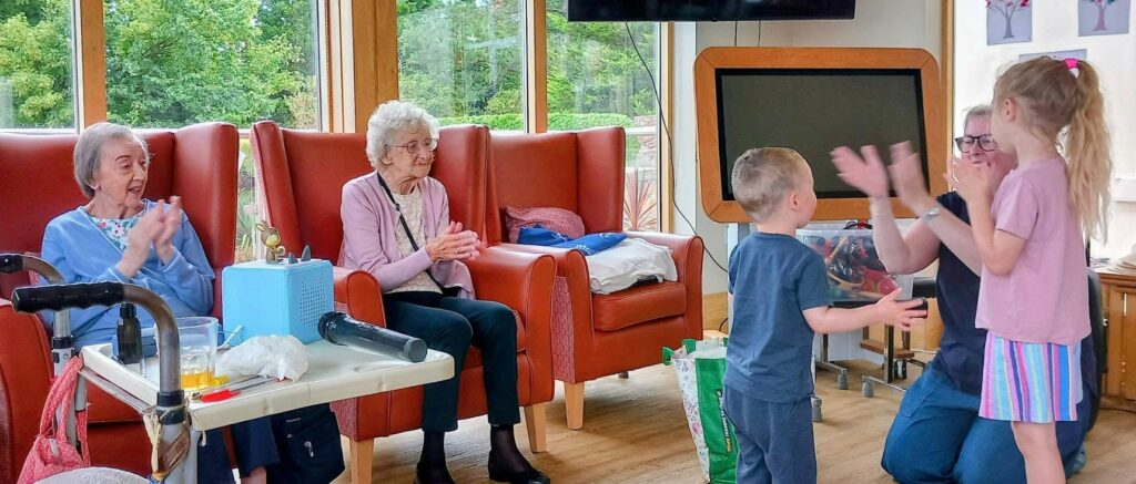 Two elderly women sit clapping, exchanging stories, as another woman kneels to play a clapping game with two young children in a bright room bathed in cheerful morning light and furnished with orange chairs.