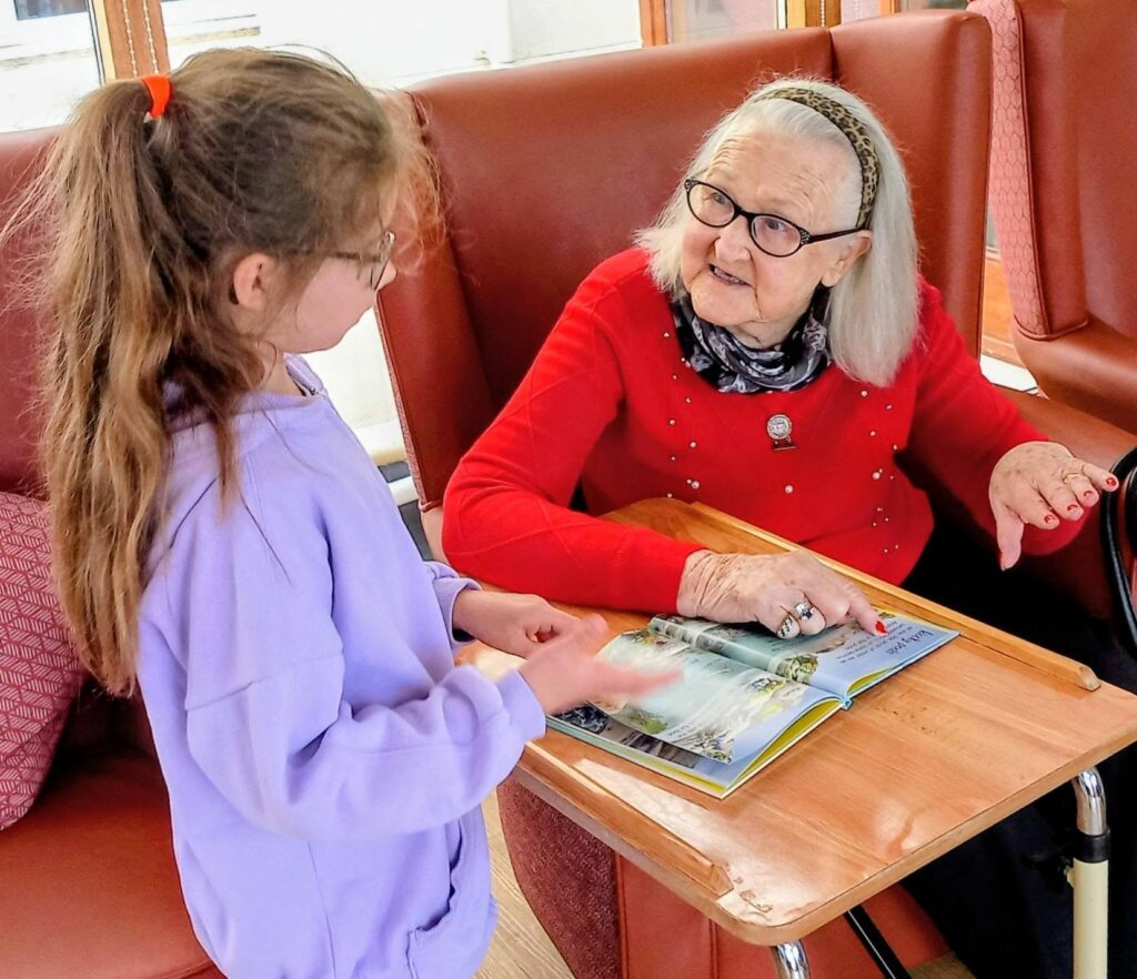 An elderly woman with grey hair and spectacles sits at a table, sharing stories with one of her youngest visitors—a young girl with long brown hair who stands holding an open magazine.