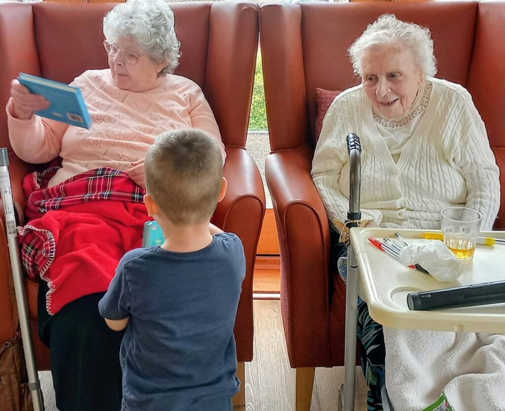 A young boy, one of the home's youngest visitors, stands between two elderly women in armchairs; one reads a card whilst the other smiles. A walking frame and tray with a drink and cutlery are nearby.