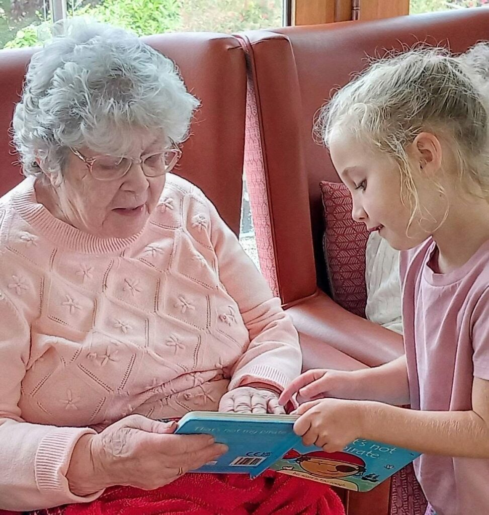 An elderly woman and a young girl, two of the library’s youngest visitors, sit on a red sofa reading a colourful book. The woman wears a light pink jumper and glasses whilst the girl points at something on the page.