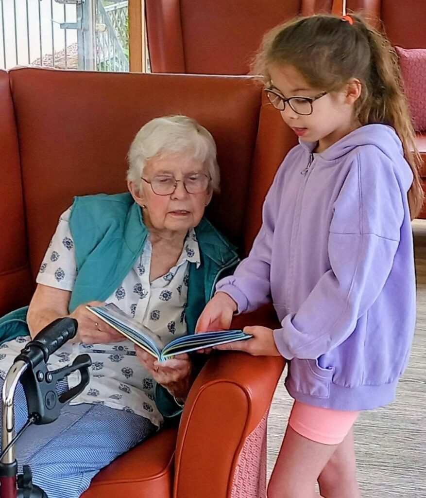 An elderly woman reads stories from a book whilst a young girl, one of the youngest visitors, stands next to her, both engrossed in the pages.