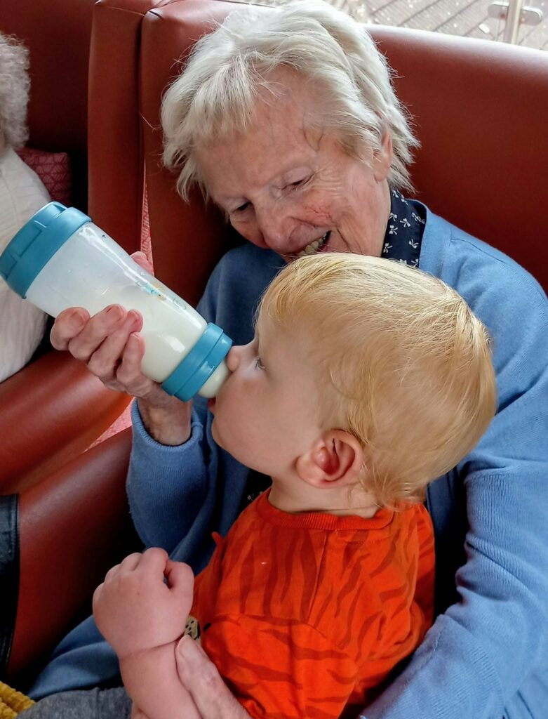 An elderly woman in a blue jumper smiles as she feeds one of her youngest visitors, a blonde child in an orange top. They sit together on a red chair, sharing peaceful moments and soft stories.