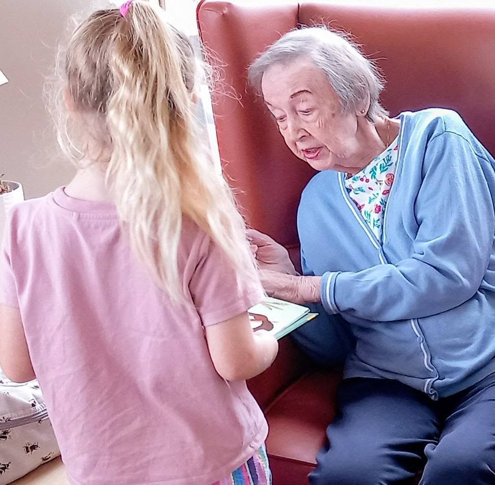 An elderly woman in a blue cardigan sits in an armchair, listening as her youngest visitor, a girl with long blonde hair, shares stories from the card she holds.
