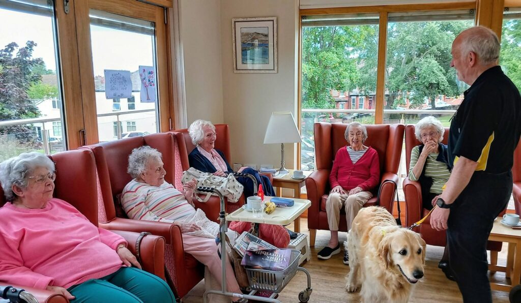 A group of elderly people sit in armchairs in a bright room, watching Gwyll and Tim from Therapy Dogs Nationwide with a golden retriever. Windows, a walking frame, and assorted items are on a small table nearby.