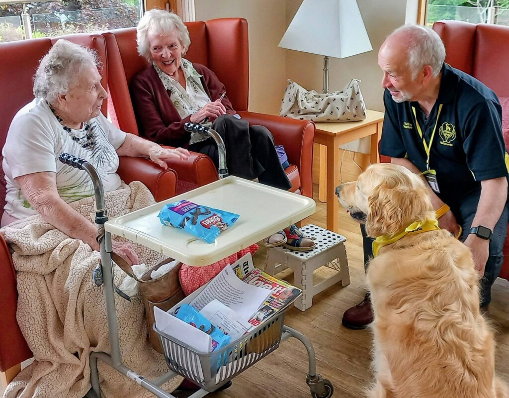 Two elderly women sit in armchairs, smiling as they engage with a man and Gwyll, a therapy dog from Therapy Dogs Nationwide. Between them is a small trolley with snacks and magazines in the bright, well-lit room.