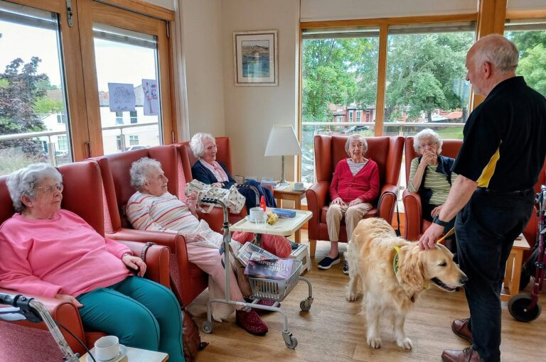 A group of elderly women sit in armchairs in a bright room, while a man stands holding a golden retriever from Therapy Dogs Nationwide on its lead.