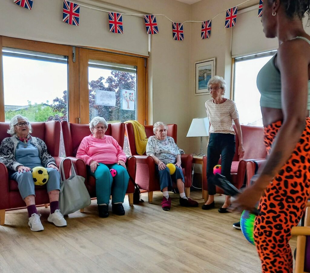 Four older women have a lively session with Carla, three seated holding balls and one standing, participating in movement exercises. Colourful leggings and Union Jack bunting brighten the sunny room with large windows.