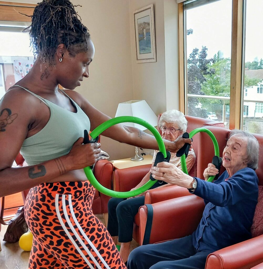 Carla leads two elderly women in enjoyable movement exercises with green fitness rings as they sit in armchairs in a bright, airy lounge.