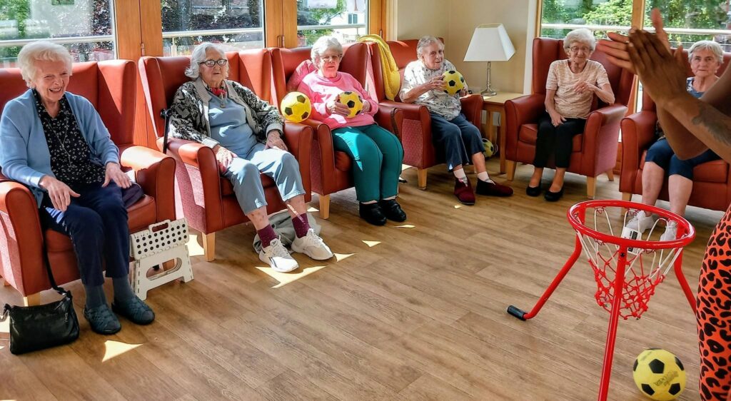 Six elderly women have an enjoyable session with Carla, moving about as they toss yellow footballs towards a small basketball hoop set up on the wooden floor.