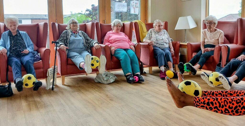 Six elderly women, including Carla, enjoy a lively activity as they sit in chairs and raise their legs, balancing small yellow footballs on their feet in a bright room.