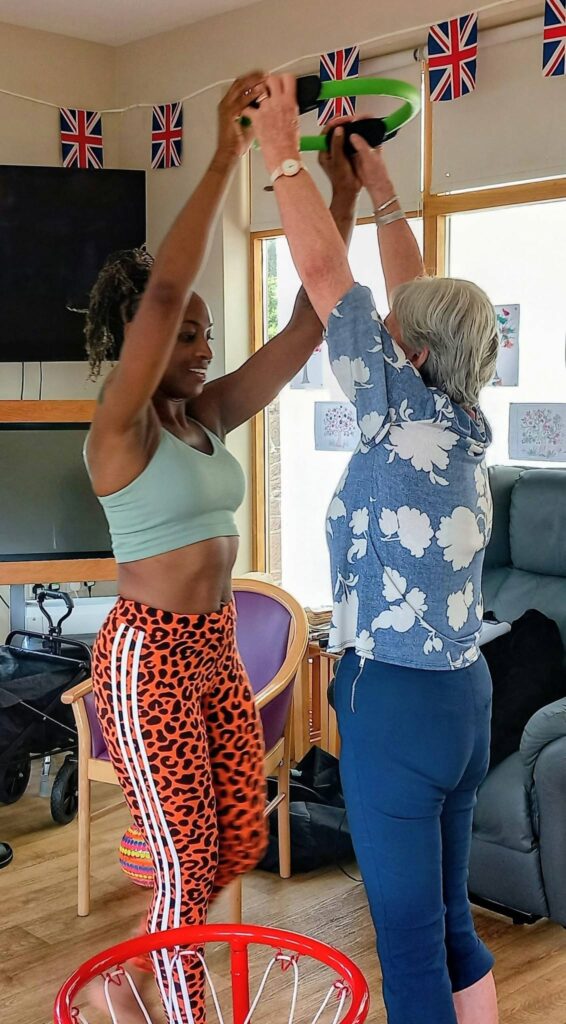 Two women take part in a lively movement session with Carla indoors, each raising a green ring above their heads. Union Jack flags adorn the background, with chairs and a walking frame close by.