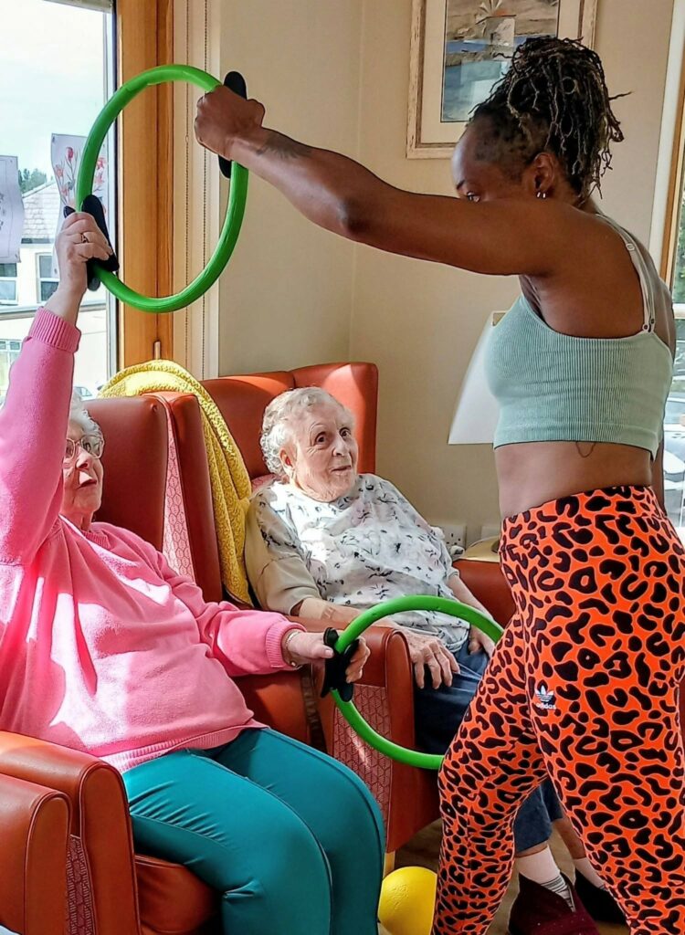 Two elderly women take part in a lively movement session with Carla, seated in armchairs holding exercise rings as she leads them in a well-lit room.