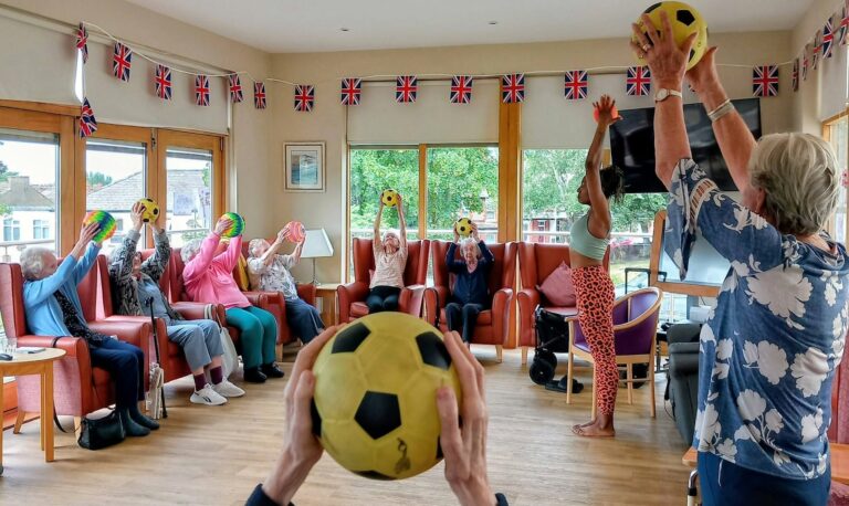 A group of older adults sit in a circle indoors, following an instructor whilst lifting yellow and black balls overhead. Union Jack flags hang in the room, and large windows reveal greenery outside.