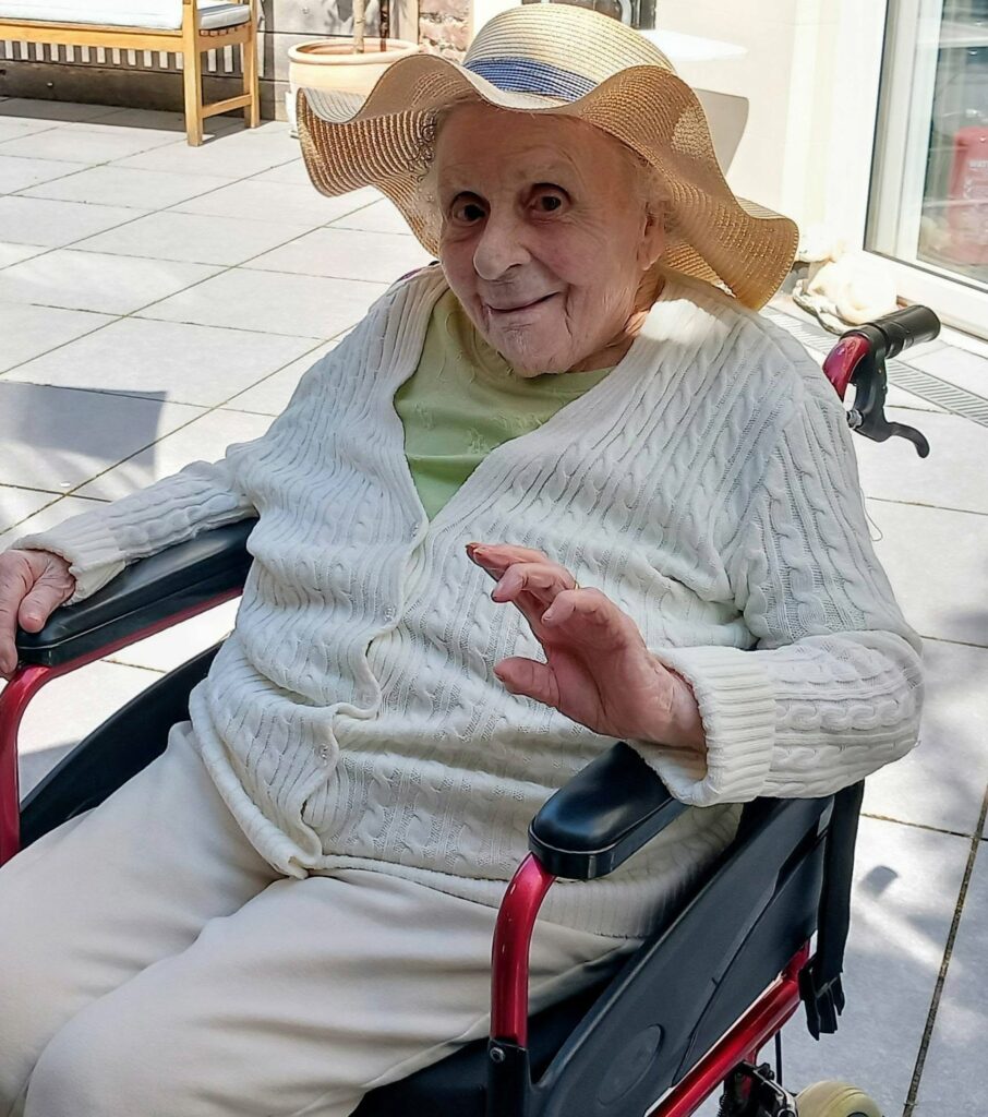 Elderly person in a sun hat and white jumper sits in a wheelchair outdoors, smiling and waving in the sunshine, with patio slabs and a wooden bench in the background.