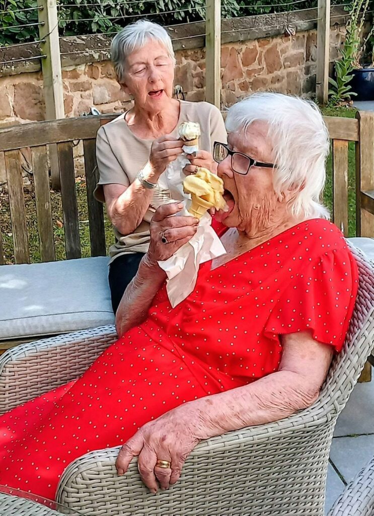 Two elderly women sit outside in a sunny garden on garden furniture, each enjoying an ice cream cone. The woman in front wears a red dress with white spots; the other, in beige, smiles as they enjoy the sunshine together.