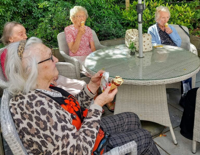 Four elderly women sit around a round wicker table outdoors, enjoying ice cream cones. Green plants appear in the background.