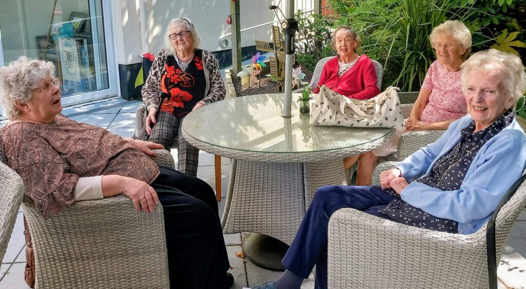 Five elderly women sit around a glass patio table outdoors, enjoying pleasant company and conversation, surrounded by lush garden plants with a building in the background.