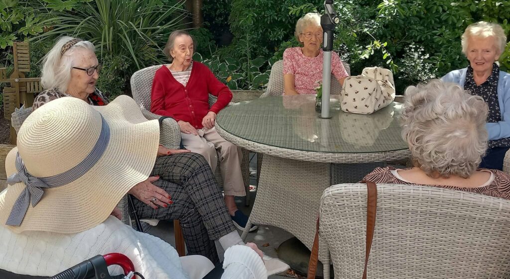 Six elderly women sit round a circular glass table in the garden, enjoying sunshine and chatting. One wears a large sunhat and uses a walking frame. A light bag sits on the table amongst the greenery.