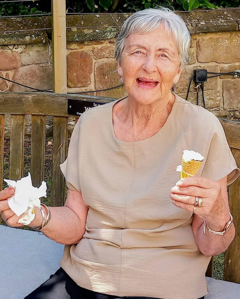 An elderly woman with short grey hair sits on an outdoor bench, smiling in the sunshine as she enjoys an ice cream. A stone wall and verdant garden greenery provide the backdrop to this joyful scene.
