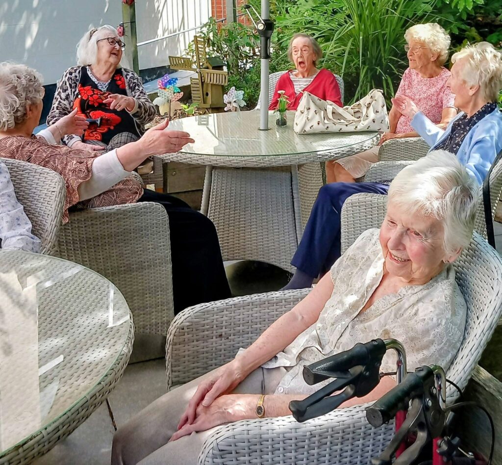 A group of elderly women sit around outdoor tables, chatting and laughing in the sunshine. One woman in the foreground smiles next to her walking frame, enjoying the fresh air and greenery in the garden.
