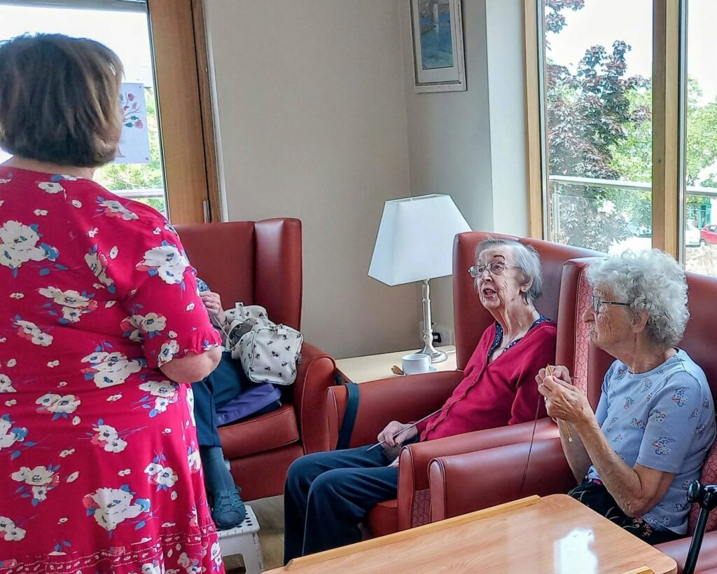 Three women meet for Knit and Natter; Heather stands as two elderly ladies sit in red chairs. Sunlight streams through the windows, brightening a cosy Thursday with table, lamp, and greenery visible outside.