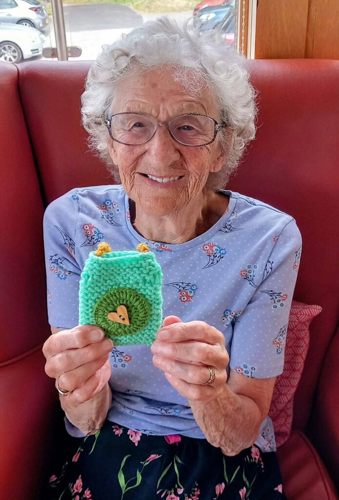 Heather, an elderly woman with grey hair and glasses, sits on a red sofa at Knit and Natter Thursday, smiling as she holds a small green knitted item decorated with a heart-shaped button.