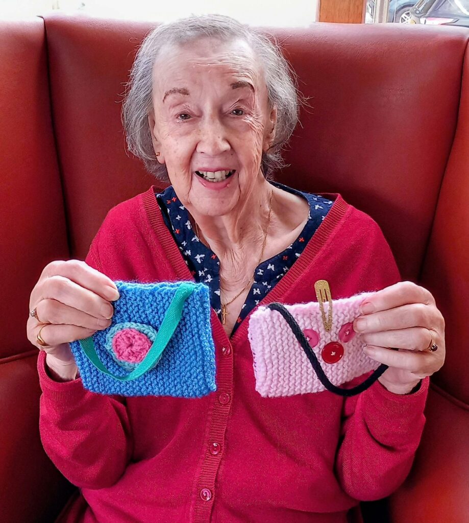 Heather smiles on Thursday at Knit and Natter, seated on a red chair as she proudly holds two small knitted purses—one blue with a pink circle, the other pink with red buttons.