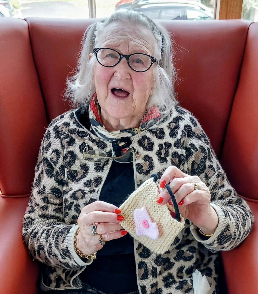 Elderly woman wearing glasses and a leopard print cardigan sits in a red chair, smiling as she holds a pink and white knitted item—possibly made at Thursday’s Knit and Natter with Heather.