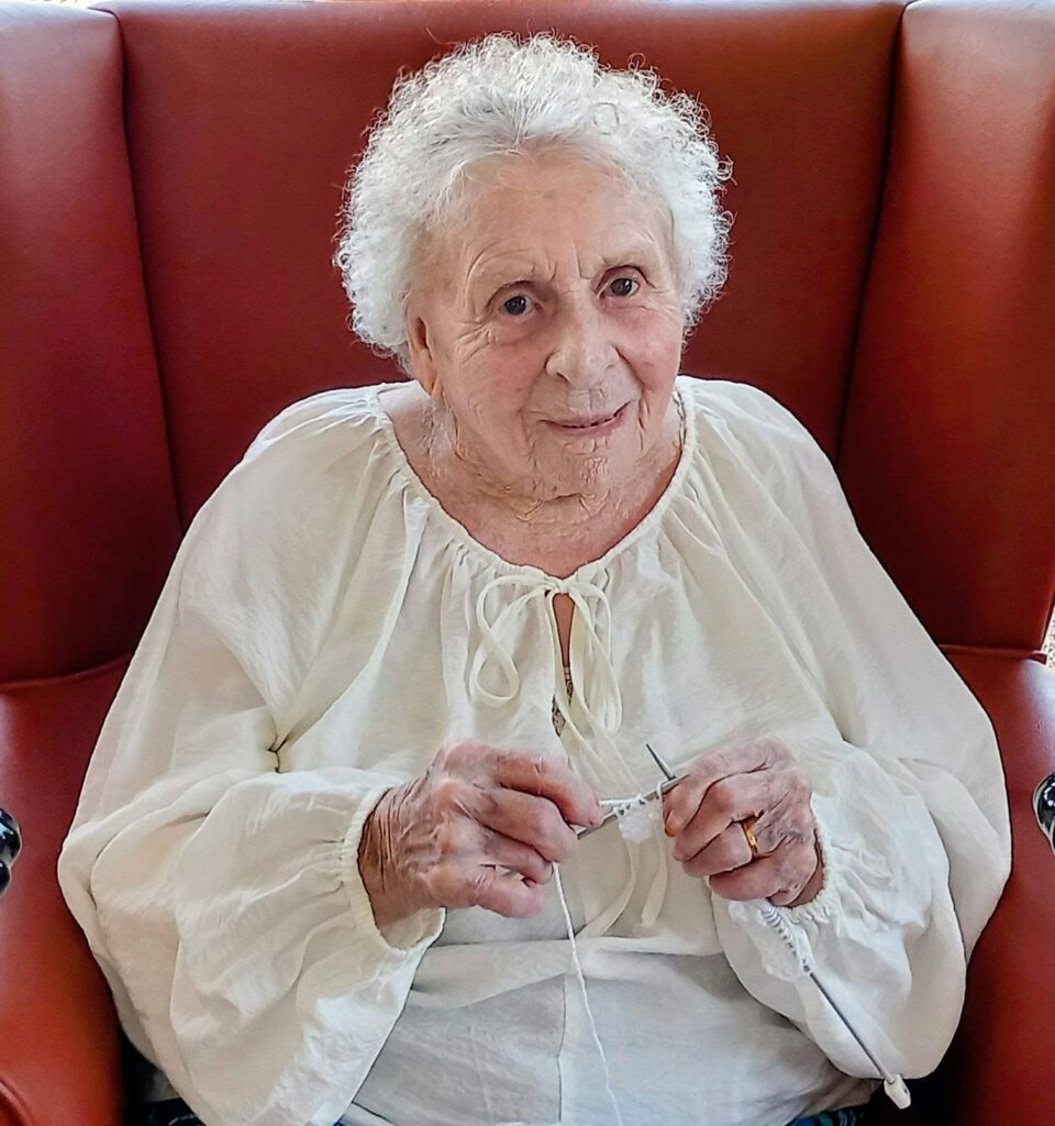 Heather, a familiar face at Knit and Natter each Thursday, sits in a red chair wearing her white blouse, holding knitting needles and wool, gazing warmly at the camera.