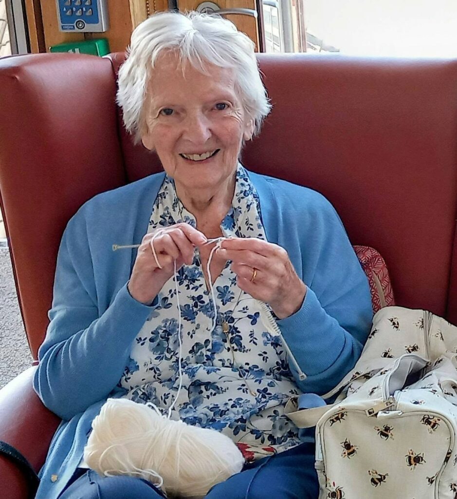 An older woman with short white hair smiles in a red chair at Knit and Natter, holding a crochet hook and white wool. She wears a blue floral blouse, blue cardigan, and has a bee-patterned bag close by.