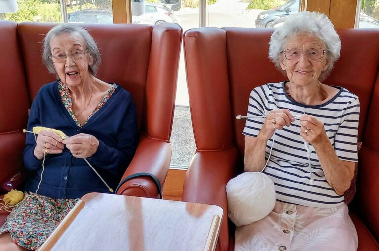 Two elderly women sit together in red armchairs, smiling as they knit with yellow and white wool at the Thursday Knit group, by a window in a brightly lit room.