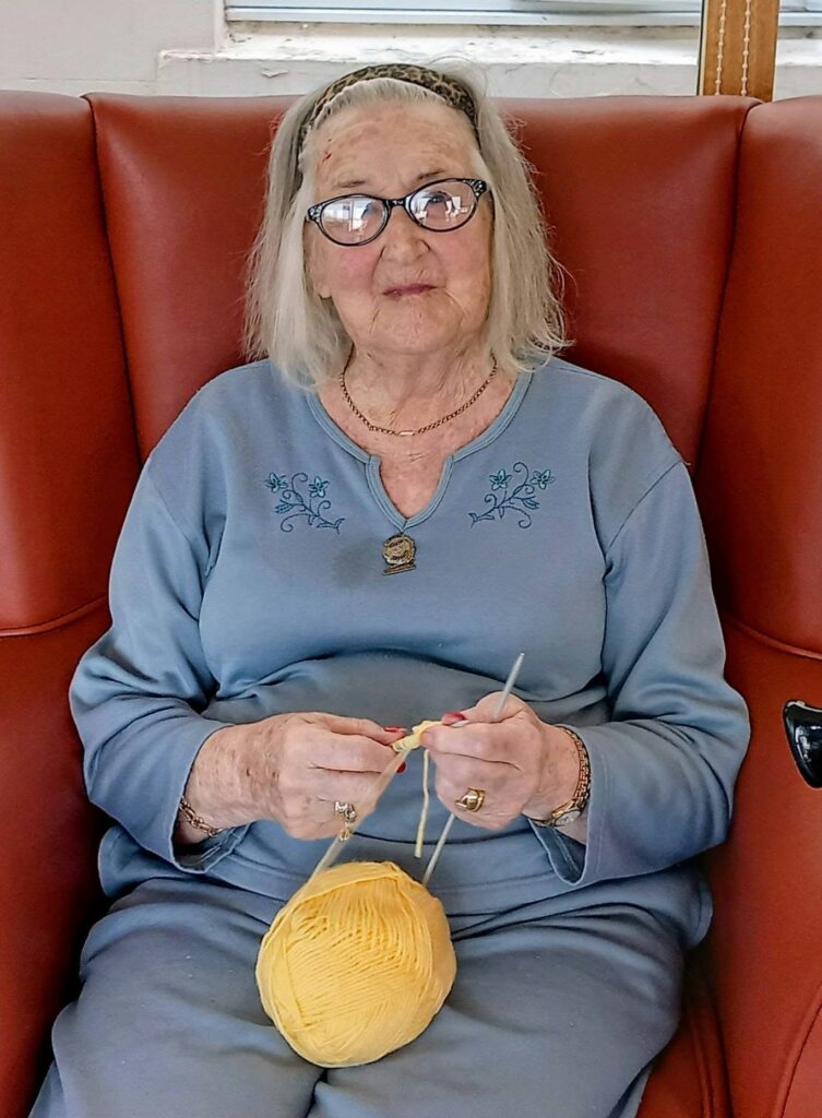 Heather, an elderly woman with spectacles, sits in a red armchair dressed in a blue outfit, knitting with yellow wool—prepared for Knit and Natter this Thursday.