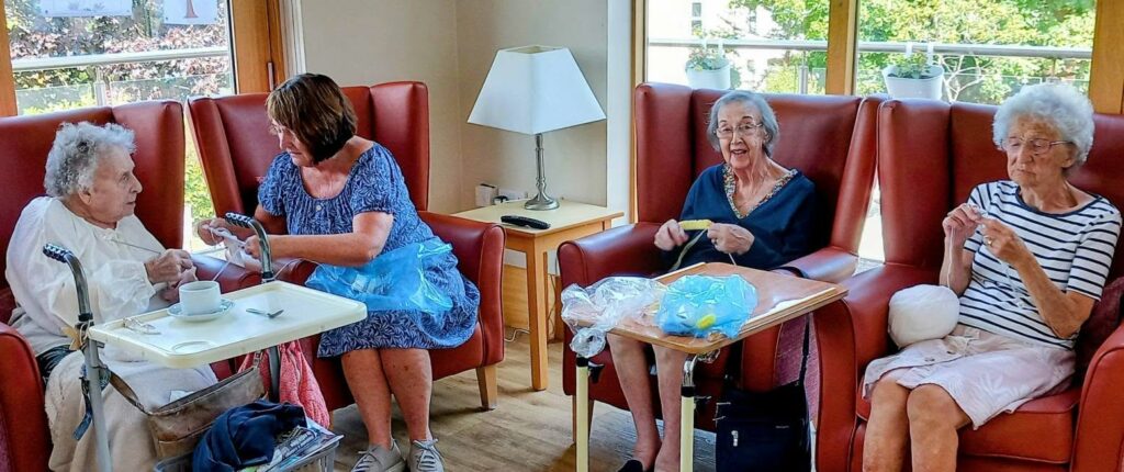 Four women gather for Knit and Natter on Thursday, sitting by a window; three are knitting while Heather in a blue dress assists another. A table with a teacup and wool sits nearby.