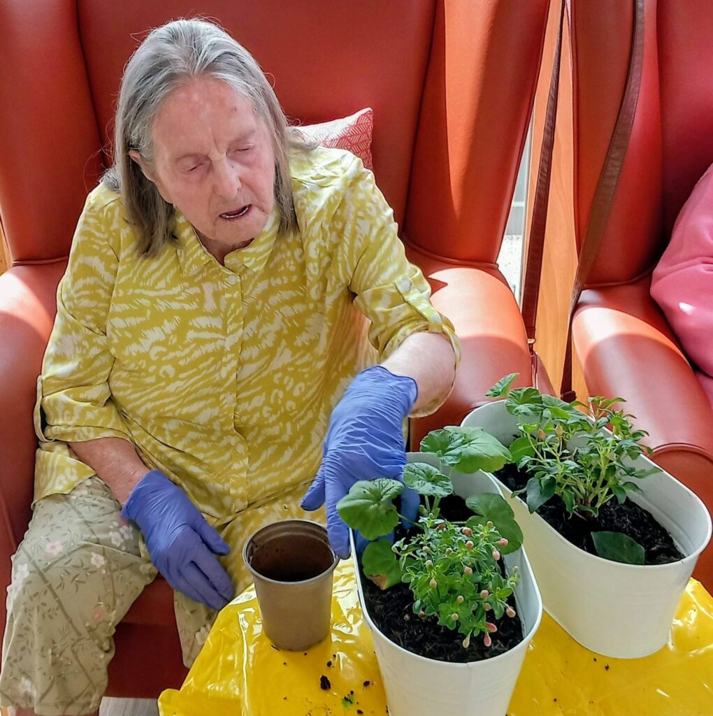 An elderly woman in a yellow shirt and blue gloves sits on a red chair at Lear House, looking after potted plants on a table covered with yellow cloth during balcony gardening.
