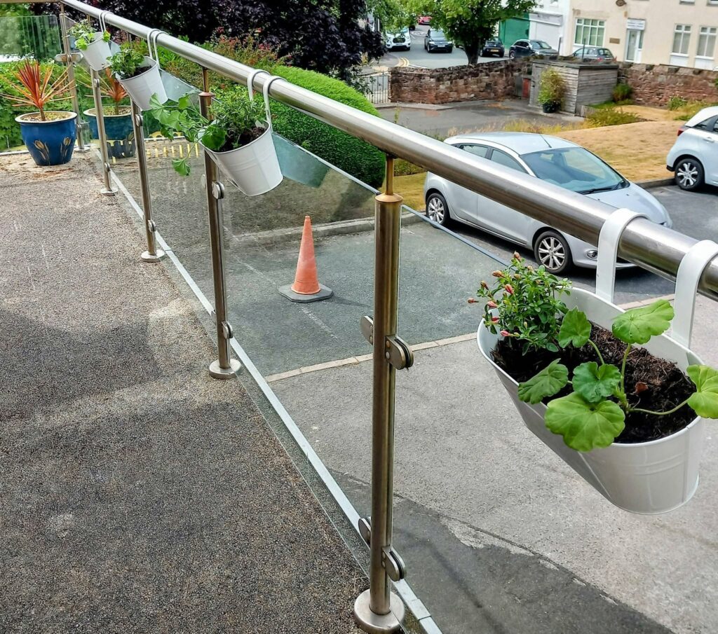White planters with green plants hang from a metal and glass balcony railing at Lear House, displaying vibrant balcony gardening. In the background, a car park with vehicles, a traffic cone, and greenery is visible.