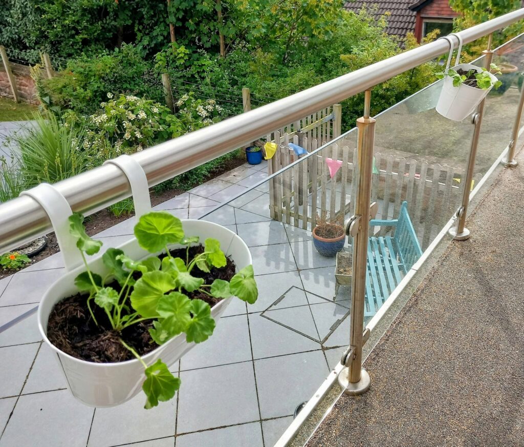 Two white hanging pots with green plants brighten the Lear House balcony railing, ideal for balcony gardening overlooking a tiled patio with a wooden bench and verdant garden.