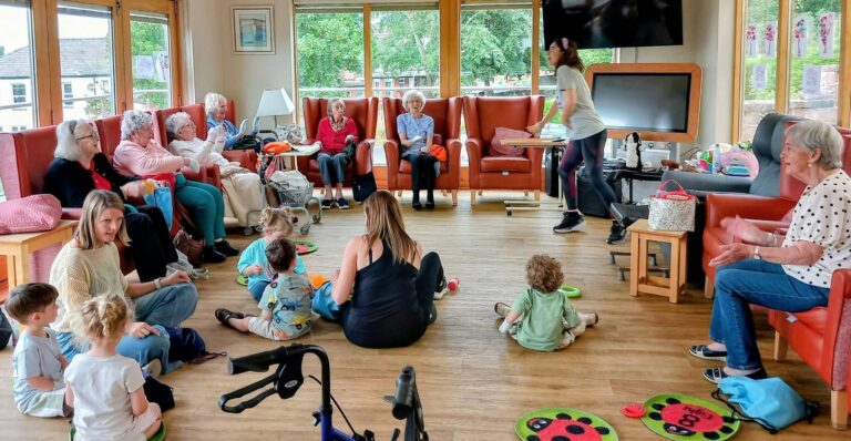 A group of elderly people and young children gather in a bright, spacious room for Popcat's Music Time, seated in a circle as they enjoy music together. A carer stands nearby, making sure everyone has an enjoyable afternoon.