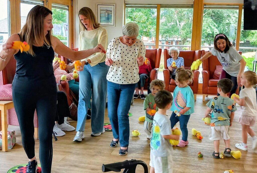 Older adults and young children enjoy Popcat's Music Time indoors, holding yellow rubber ducks. Some do the Hokey Cokey while others look on. Large windows reveal green scenery outside.