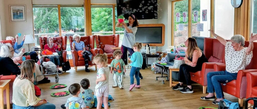Children and adults gather in a bright room for Popcat’s Music Time, surrounded by colourful props. Some sit as children in the centre sing along and dance to the Hokey Cokey, filling the space with music and laughter.