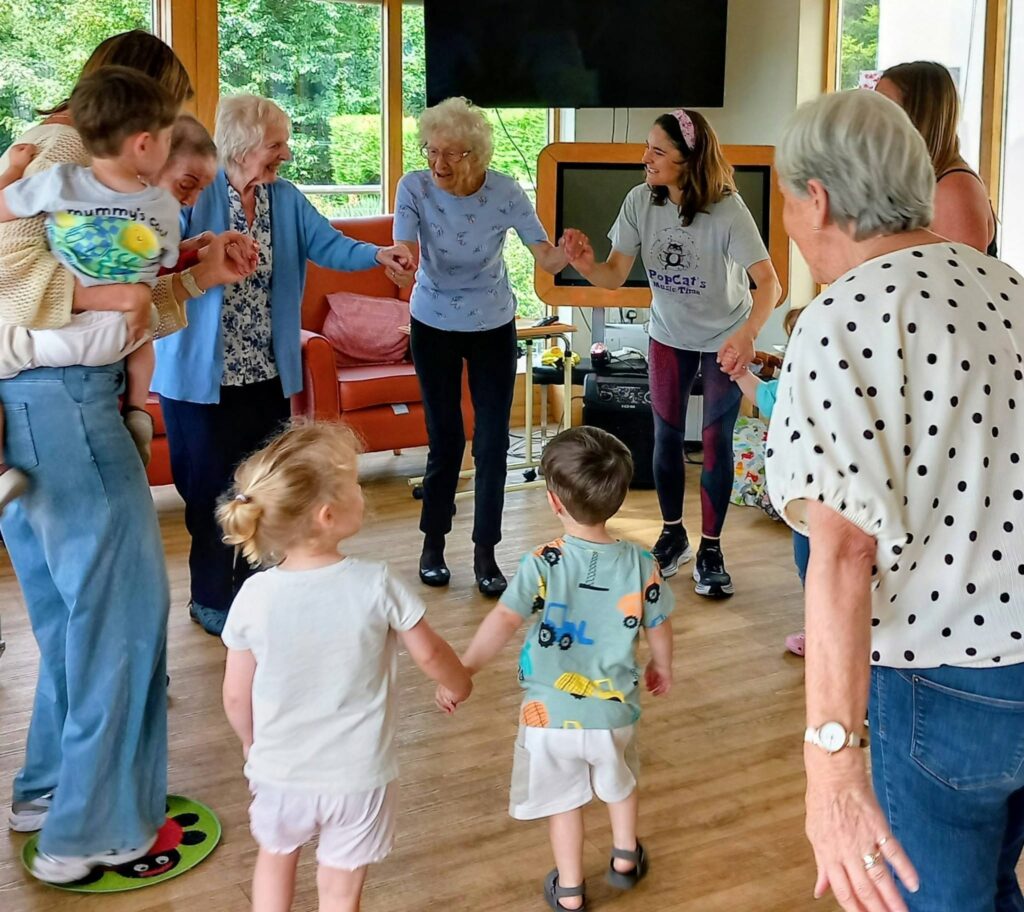 Children and elderly adults stand in a circle holding hands indoors, enjoying Popcat’s Music Time, smiling and engaging warmly as they take part in this cheerful intergenerational activity.