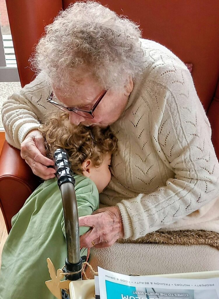 An elderly person with curly grey hair hugs and kisses a young child during a joyful afternoon, seated near a walking frame and magazine, as they enjoy Popcat's Music Time together.