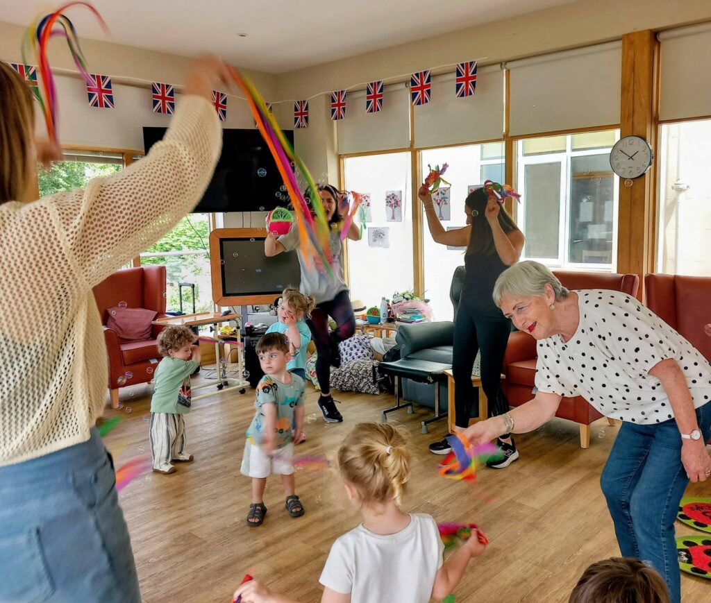 Adults and young children dance to lively music, play Hokey Cokey, and twirl colourful ribbons in a room adorned with Union Jack flags.