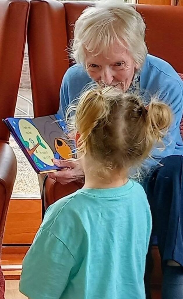 An older woman and a young girl with blonde plaits enjoy a lively afternoon indoors by a window, exploring Popcat’s Music Time picture book together.