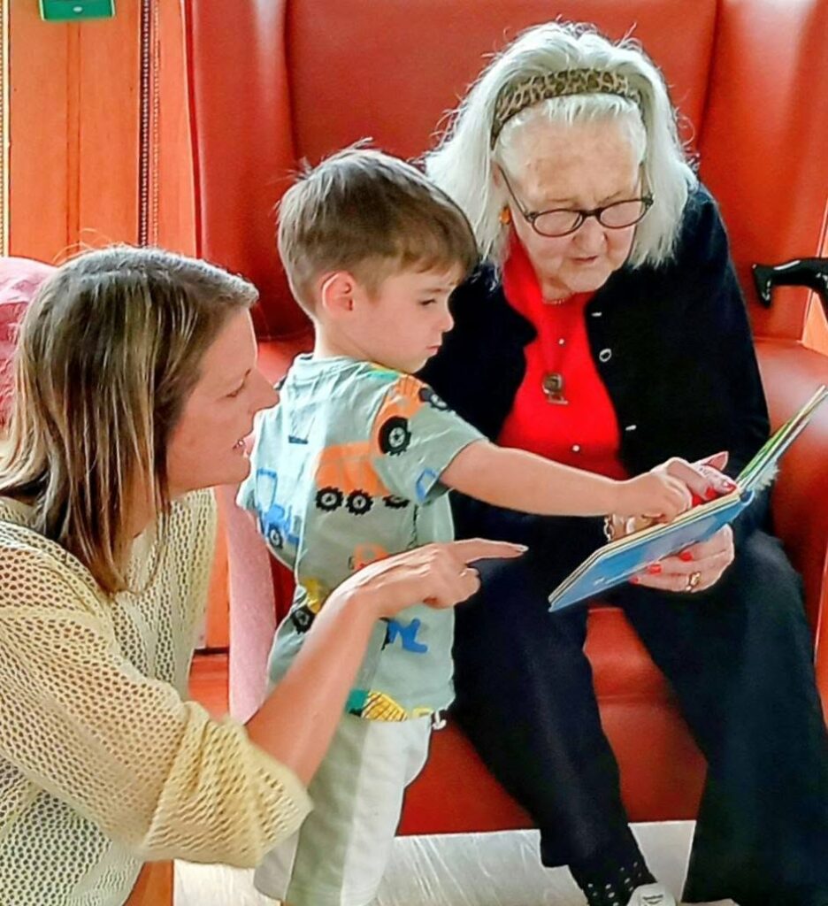 An elderly woman, a young boy, and a woman enjoy Popcat's Music Time as they look at a book together in a cosy room with warm lighting, their laughter bubbling like cheerful tunes.