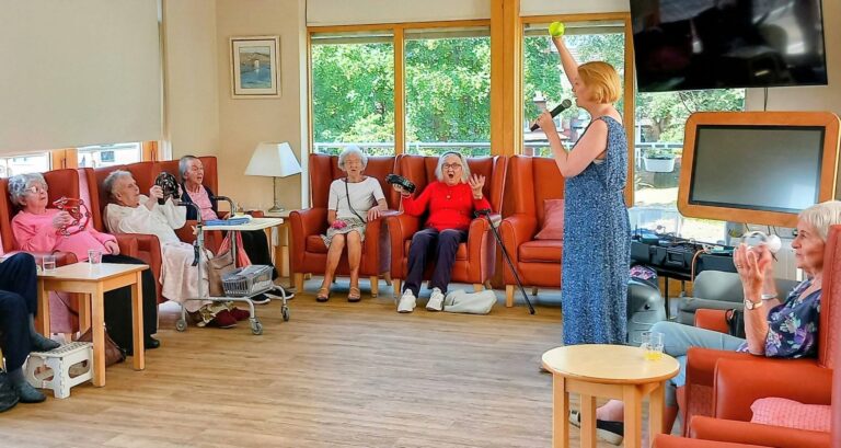 On a cheerful afternoon, Sandra Currie stands in a blue dress holding up a yellow ball as elderly women in the lounge join in enthusiastically, some clapping and others with musical instruments.