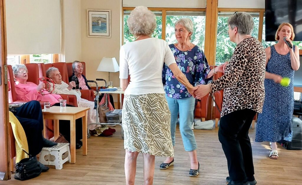 On a cheerful afternoon, Sandra Currie sings into a microphone as older women dance in the bright room, whilst others sit and watch in a laid-back atmosphere.