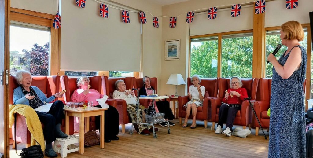 On a sunny afternoon, Sandra Currie stands at the front, speaking or singing as a group of elderly women relax in armchairs beneath Union Jacks and sunlit windows.