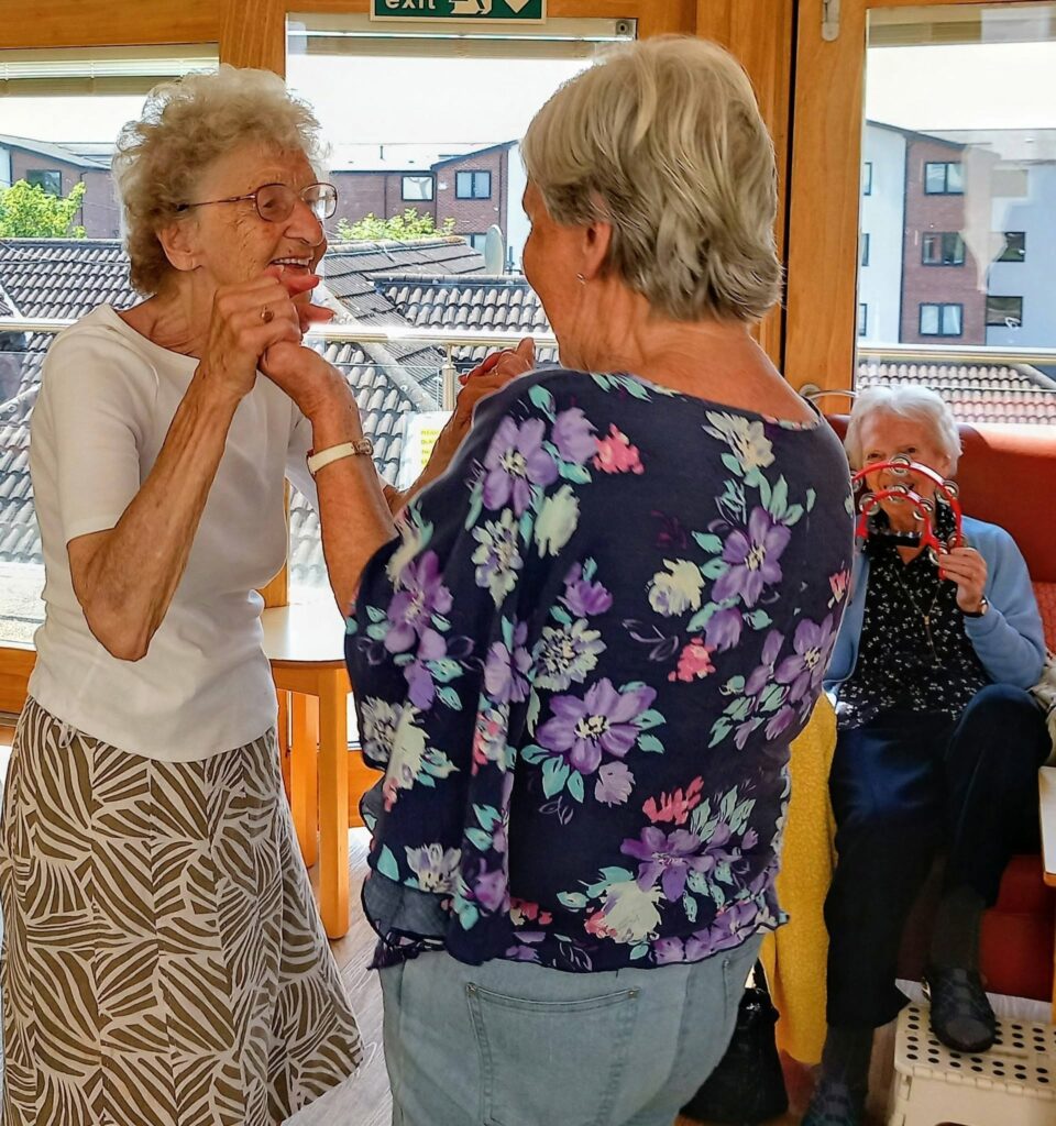 Sandra Currie captures a lovely afternoon as two elderly women stand smiling and holding hands in a sunlit room, whilst another woman plays the tambourine. Blocks of flats can be seen through the windows.