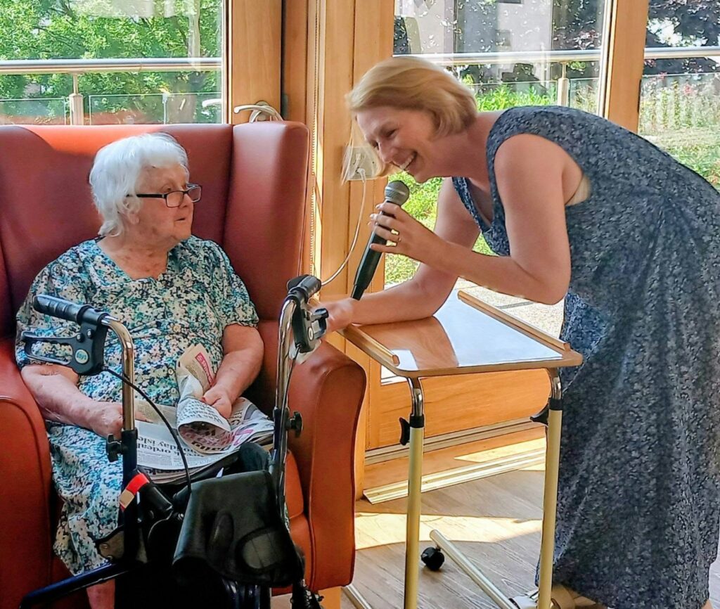 In a sunlit room, Sandra Currie smiles as she holds a microphone towards an elderly woman reading a newspaper. Nearby, a zimmer frame and tray table complete the cosy afternoon scene.