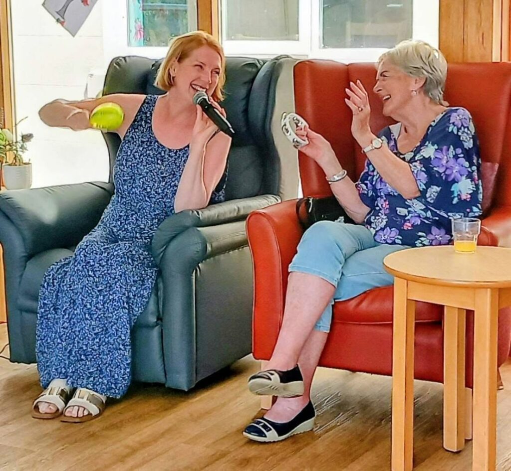 Sandra Currie enjoys a lively afternoon indoors, smiling with a microphone and yellow ball, while her friend laughs holding a tambourine. A small table with a drink rests beside them.